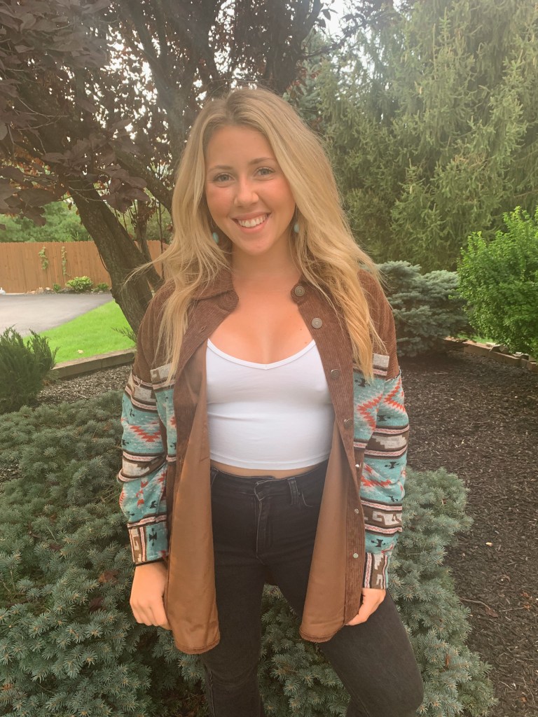 I am standing in front of a tree smiling.  I have blonde hair and blue eyes. I am wearing black jeans, a white top and a corduroy jacket